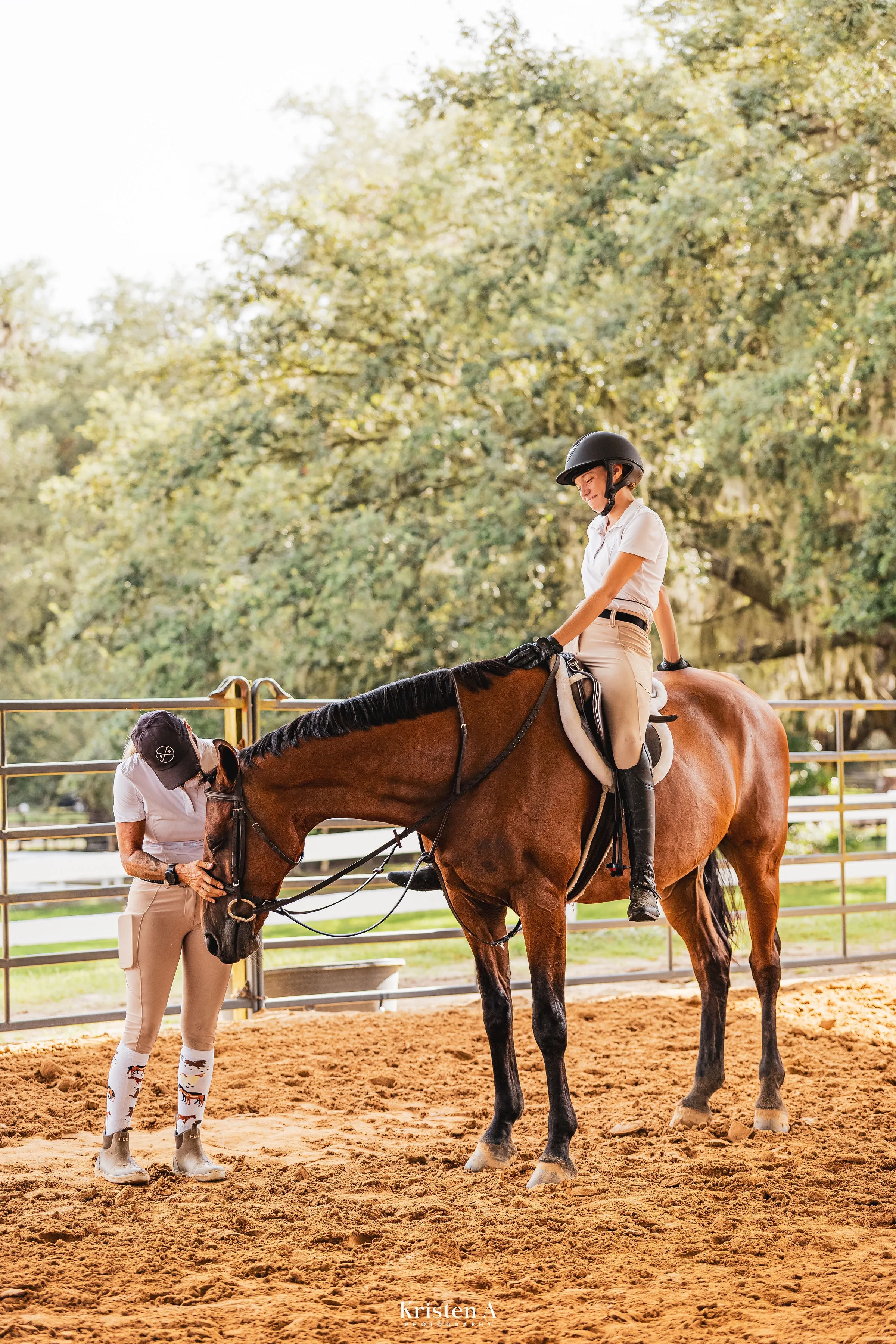 A trainer resting her hand on a bay horse’s face while a young rider pats its neck, in a sand arena under live oaks