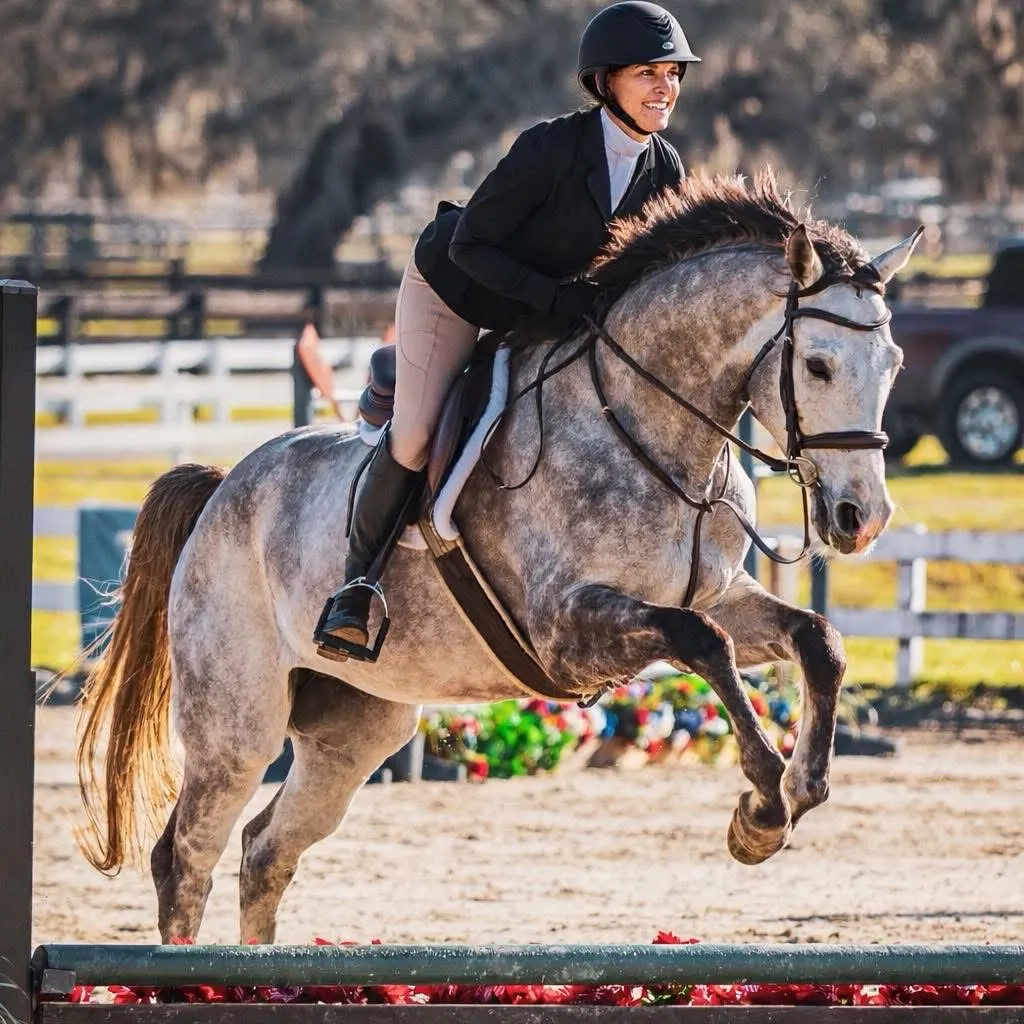 A smiling rider in a black show coat jumping a dappled grey horse over a rail