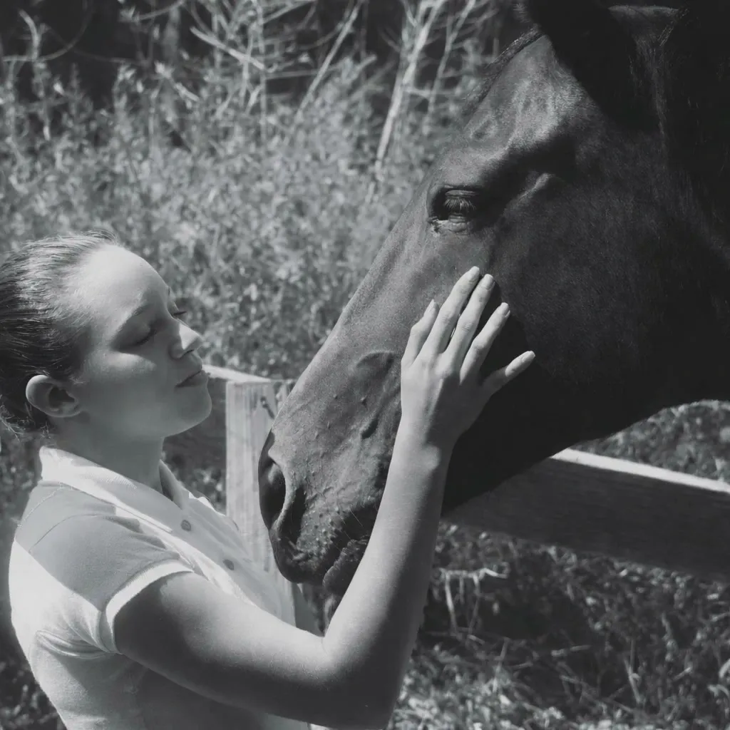 Black and white photo of a young woman resting her hand on a dark horse's face by the fence