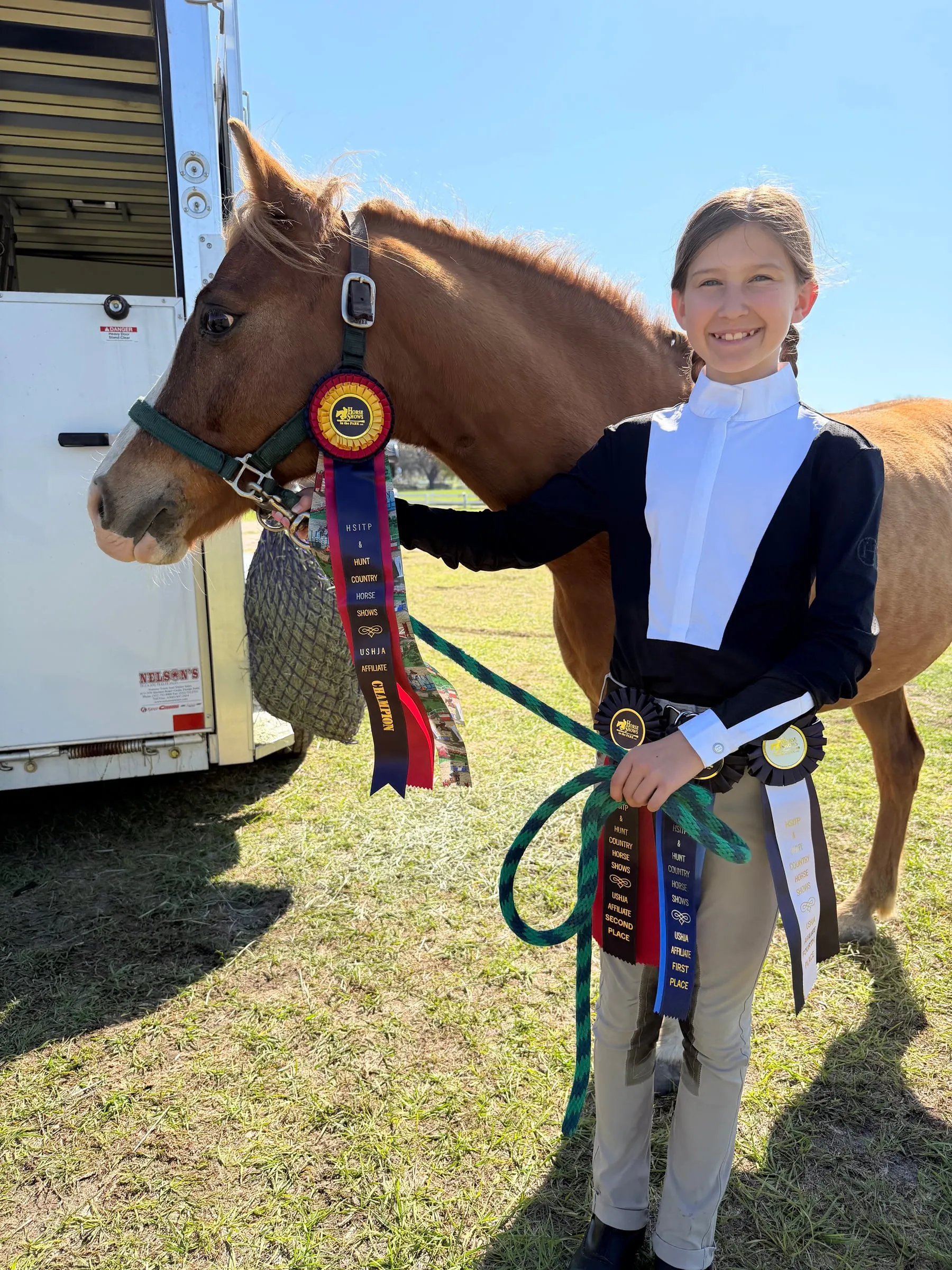 A smiling young rider beside her chestnut pony and its ribbons at the trailer